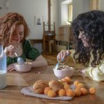 Two young women enjoying breakfast at home with cereal, croissants, and fruit around a wooden table.